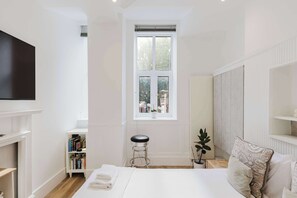 A bedroom with a large window, a bookshelf, a bedside table, and a bed with pillows. - Chic 1BD Flat in Historic Building, East London (Greater London)