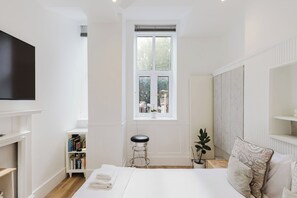 A modern bedroom with a large bed, a TV, a window, a bookshelf, and a plant. - Chic 1BD Flat in Historic Building, East London (Greater London)