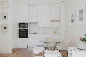 A modern kitchen with white cabinets, a glass table, and a white chair. - Chic 1BD Flat in Historic Building, East London (Greater London)