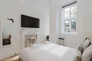 A hotel room with a large bed, a flat-screen TV mounted above a fireplace, a window with a view of trees, and a small stool.