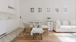 A modern, minimalist living space with a white sofa, a round glass table, and a small white stool. There are framed pictures on the wall and a potted plant on a side table.