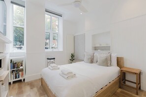 A bedroom with a large bed, a bedside table, a bookshelf, and a window with blinds. - Chic 1BD Flat in Historic Building, East London (Greater London)