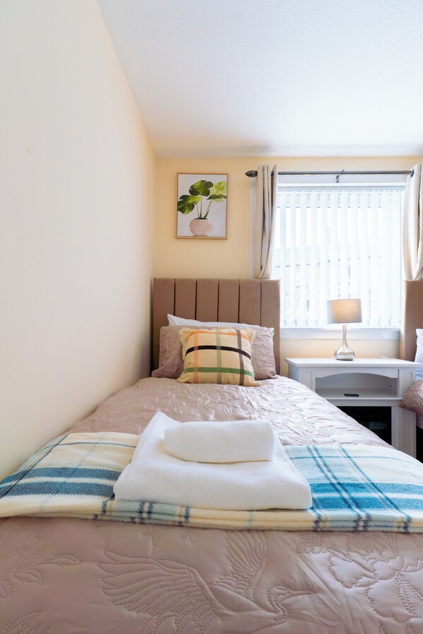 Una cama bien hecha con una manta escocesa, una toalla doblada y una almohada. Hay una mesita de noche con una lámpara, un cuadro en la pared y una ventana con cortinas.