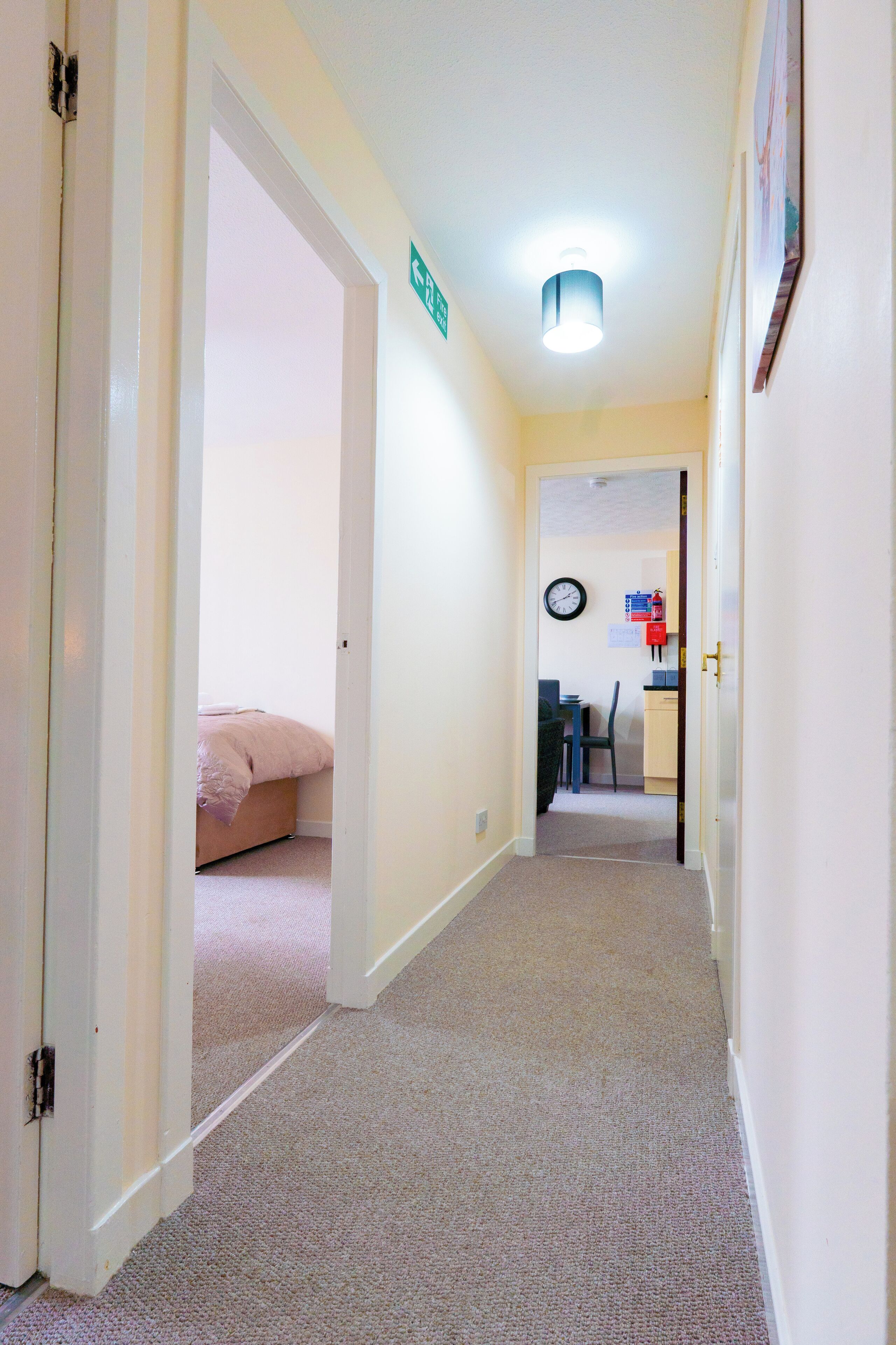 A hallway with carpeted floors, a bed in the background, a clock on the wall, and a door with a sign.