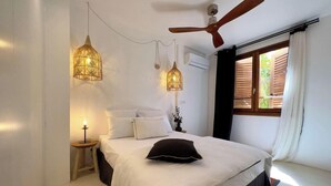Un dormitorio con cama, dos lámparas colgantes, ventilador de techo, mesita de noche con lámpara y ventana con cortinas.