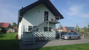 Un immeuble de deux étages doté d’un balcon, d’un escalier et d’une voiture garée devant.