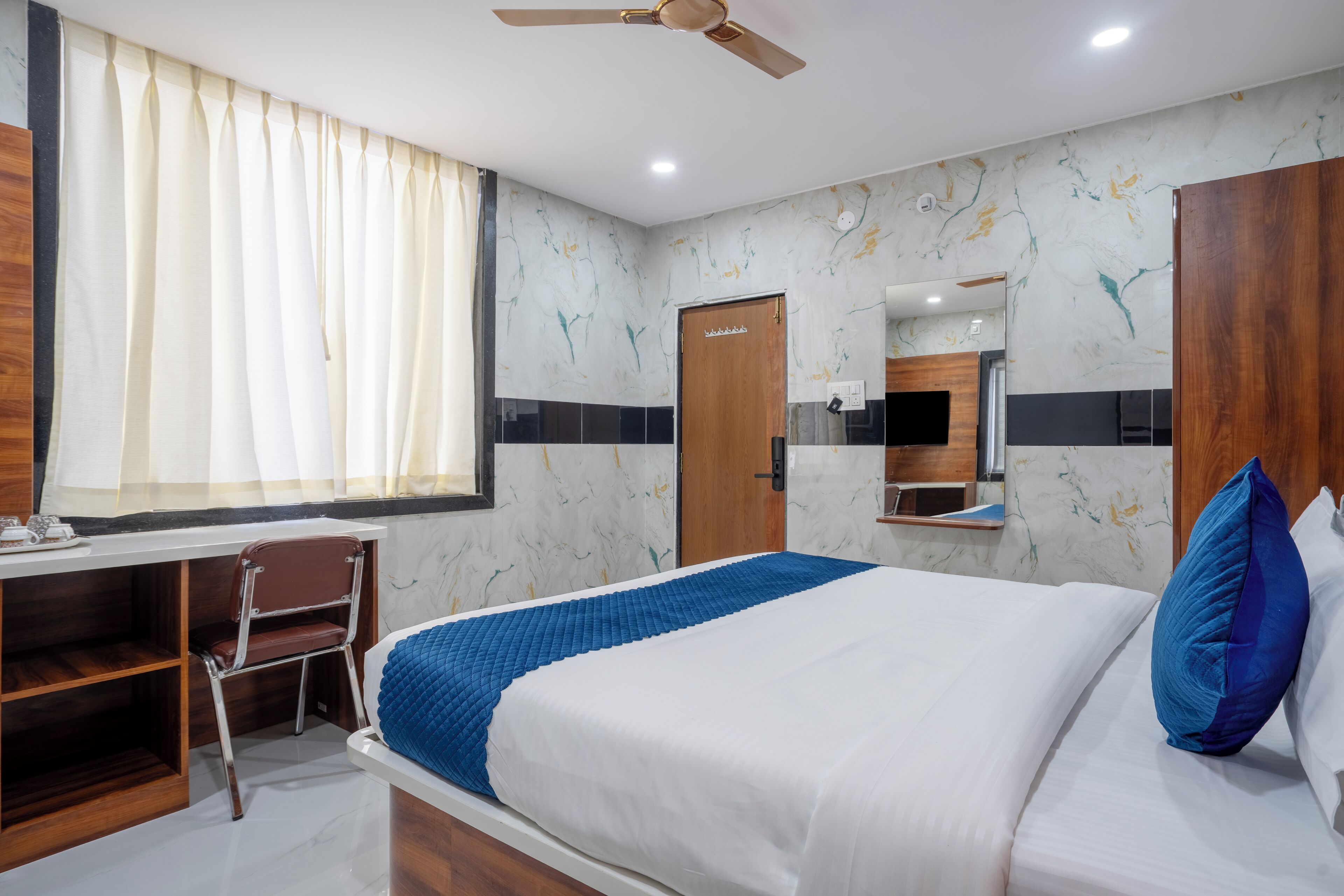 A hotel room with a bed, a desk, a chair, a ceiling fan, and a window with curtains.