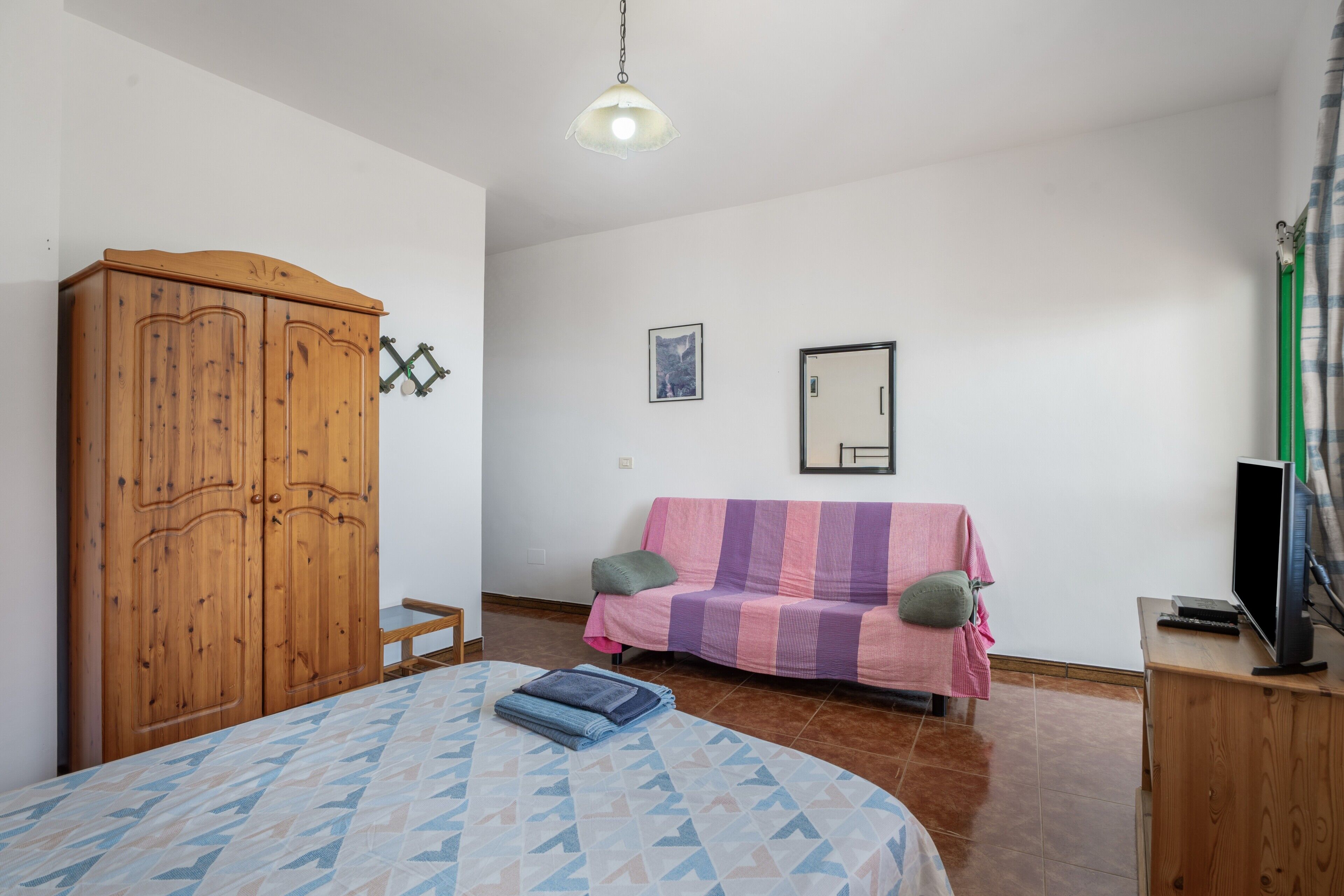 A bedroom with a bed, a wooden wardrobe, a sofa, a television, and a framed picture on the wall.