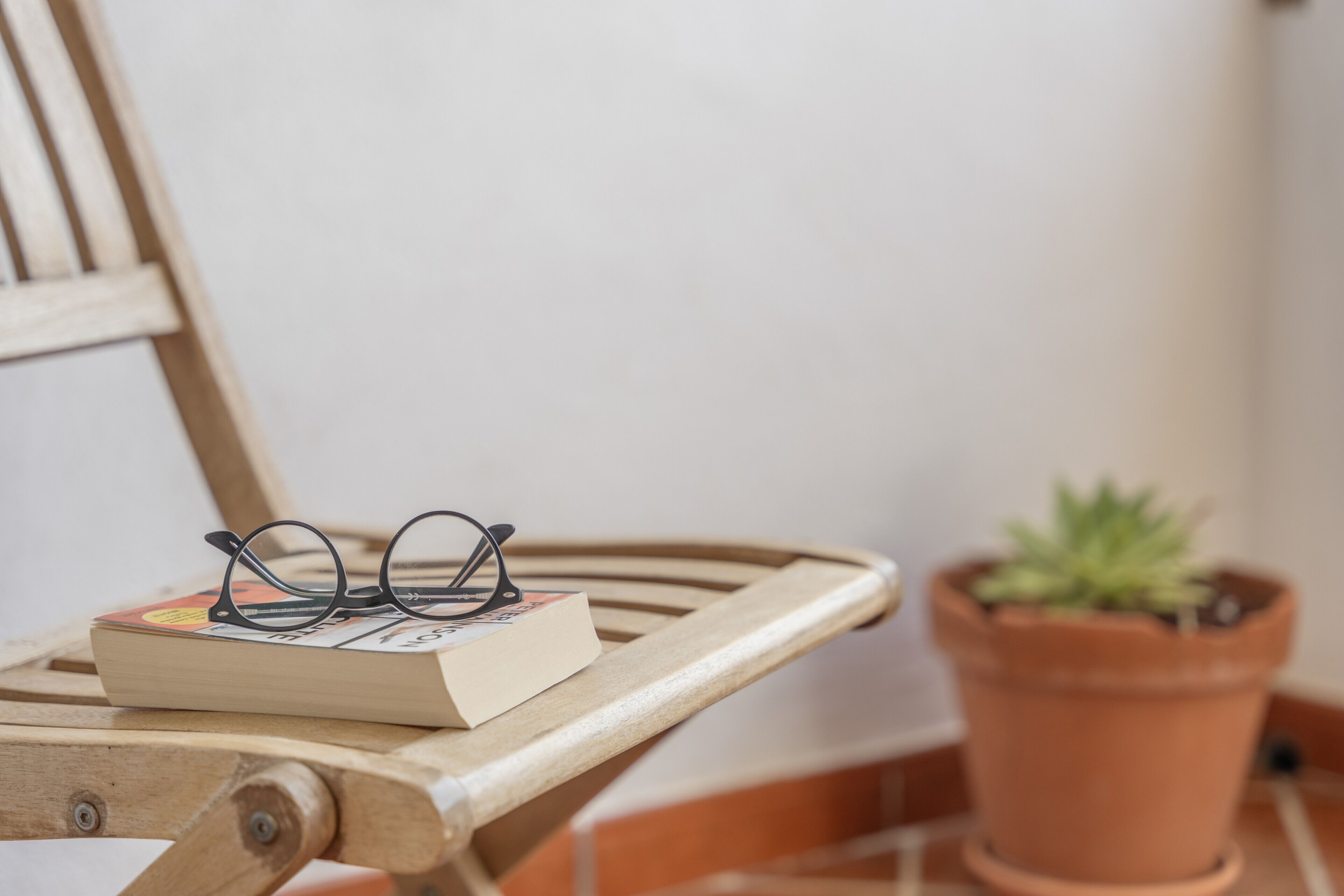 A wooden chair with a pair of glasses and a book on it, a terracotta pot with a plant, and a white wall in the background.
