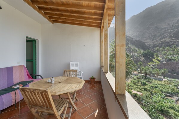 Un balcón con una mesa y sillas de madera, una puerta verde y vistas a un paisaje montañoso.