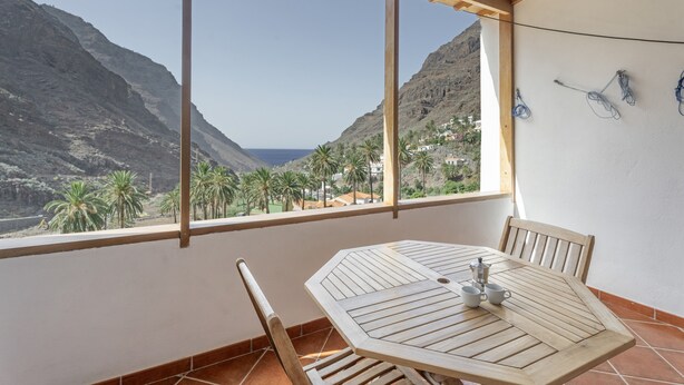 A balcony with a wooden table and chairs, a view of mountains and palm trees, and a white wall with a power outlet. - Apartment 'Nayara A' with Sea View, Balcony and Wi-Fi (Casa de la Seda)