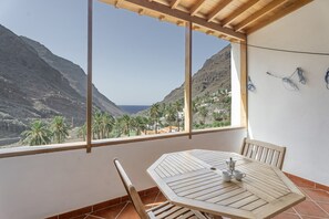 Un balcón con mesa y sillas de madera, vista a montañas y palmeras, y una pared blanca con toma de corriente.