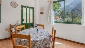 A room with a round table, chairs, a clock, a green door, a window with a view of mountains, and a water bottle on the table. - Apartment 'Apartamento Nayara B' with Sea View, Shared Terrace and Wi-Fi (Valle Gran Rey)