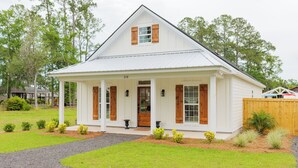 Een wit huis met één verdieping en een puntige dakkapel, nummer 218, met een veranda met kolommen, houten luiken en een kleine voortuin met beplanting.