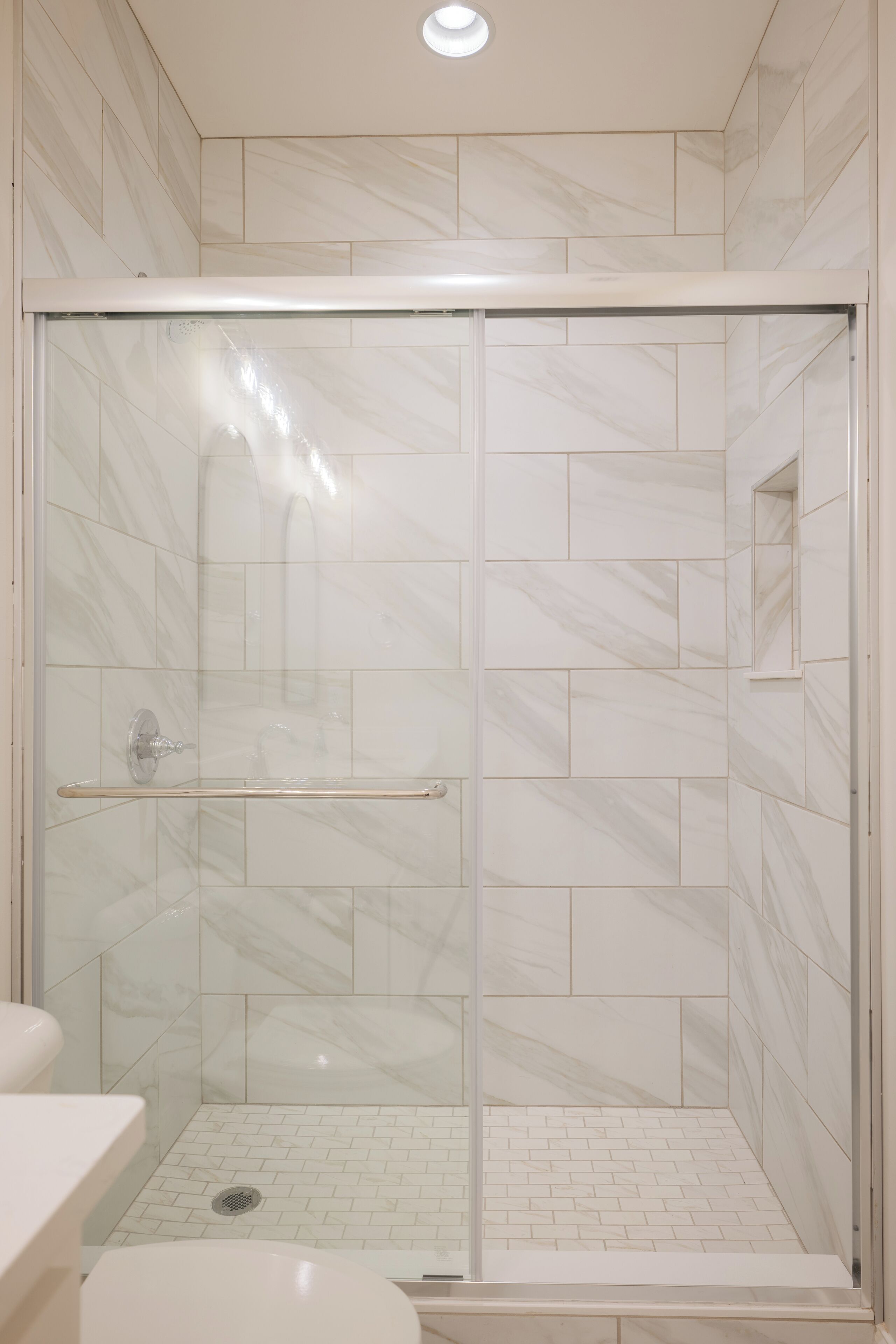 Une douche à l’italienne avec une porte en verre, des carreaux de marbre blanc et une barre d’appui.