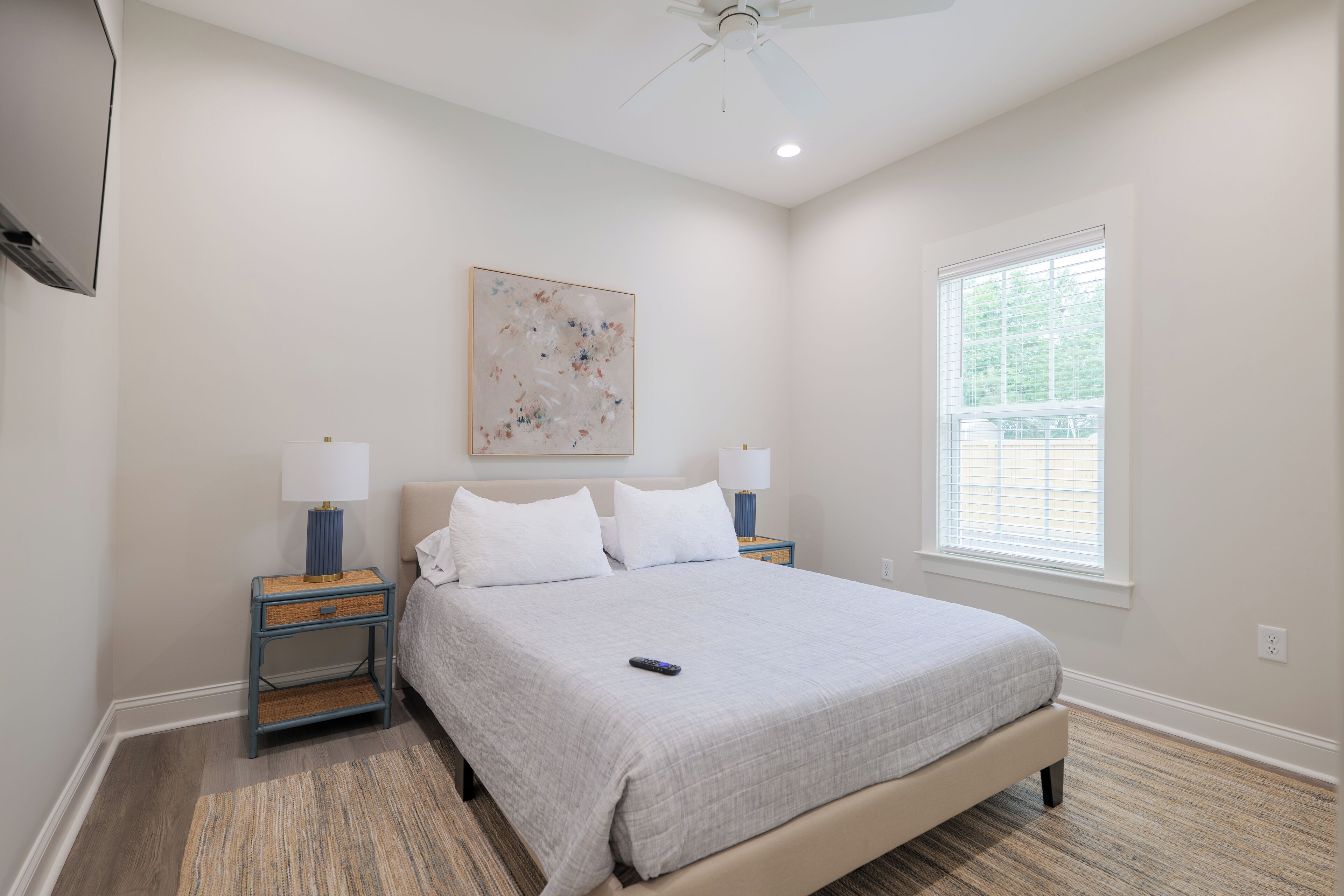 A bedroom with a bed, two bedside tables, a ceiling fan, a window, and a wall-mounted TV.