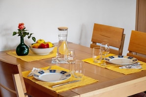 A wooden dining table set with plates, cutlery, a vase with a flower, and a bowl of fruit.
