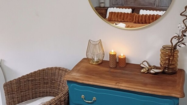 A room with a round mirror, a blue dresser with gold handles, a wicker chair, and a wooden nightstand with a lamp.