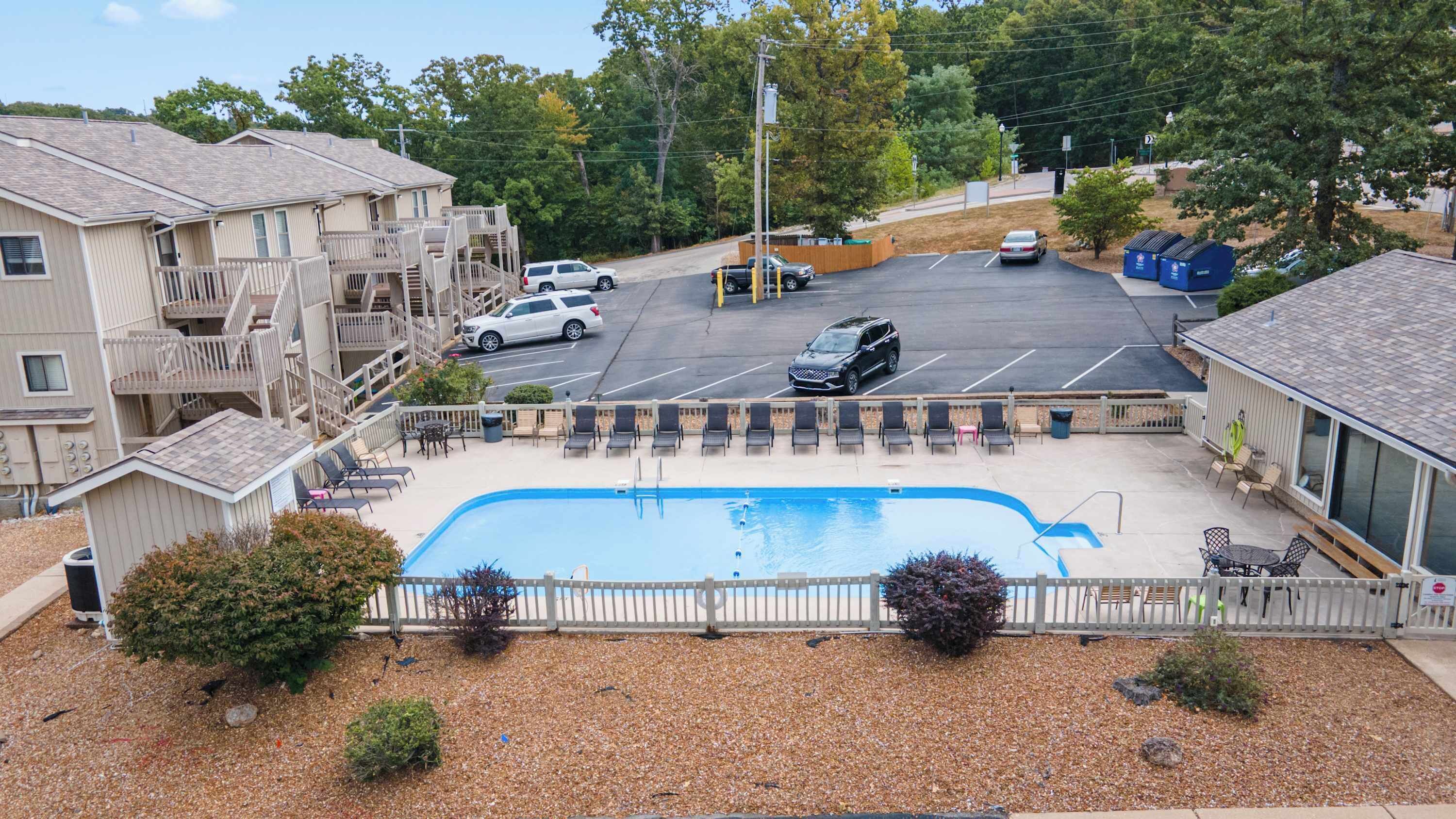 A pool area with lounge chairs, a fenced swimming pool, and a parking lot with cars.