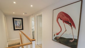 Un couloir avec un escalier en bois, un tableau encadré représentant un flamant rose et une porte blanche donnant accès à une salle de bain.