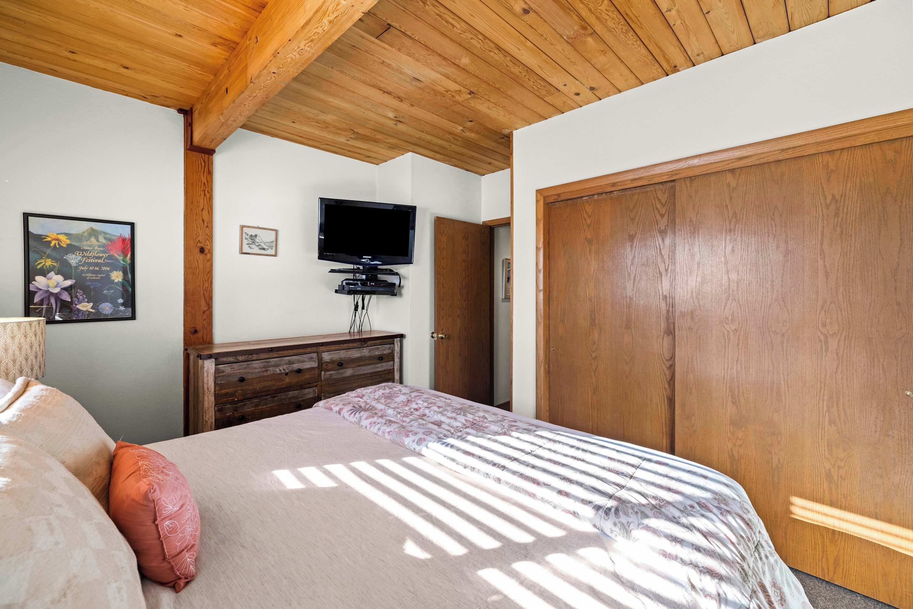 A bedroom with a wooden ceiling, a bed with a floral bedspread, a television on a stand, and a wooden wardrobe.