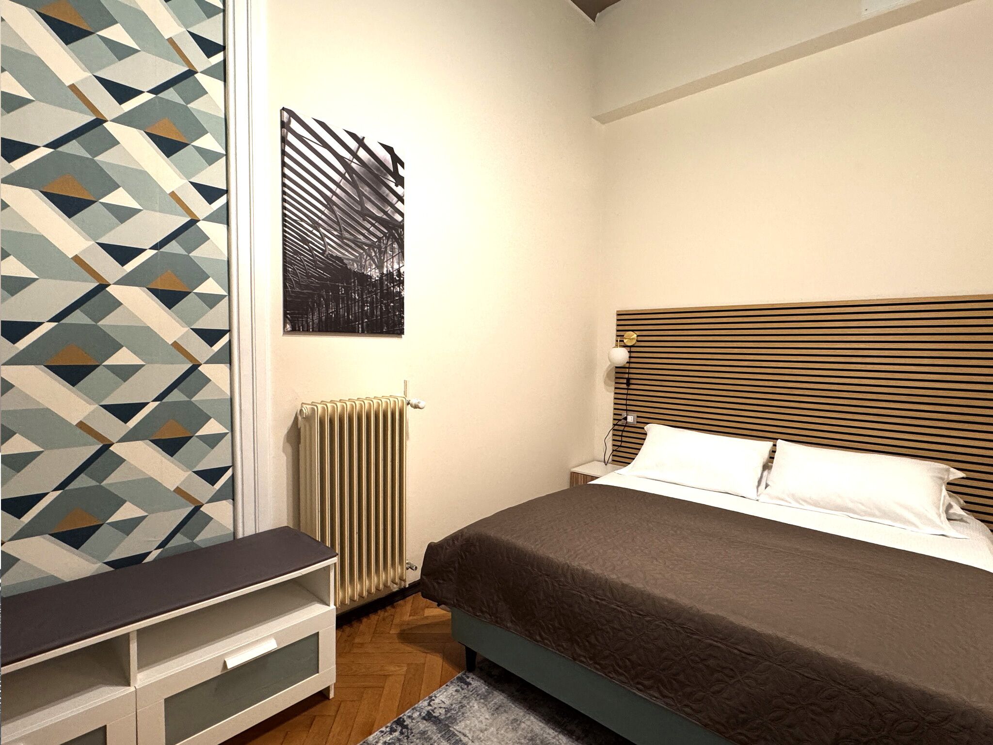 A bedroom with a bed, bedside table, radiator, and a geometric-patterned wall.