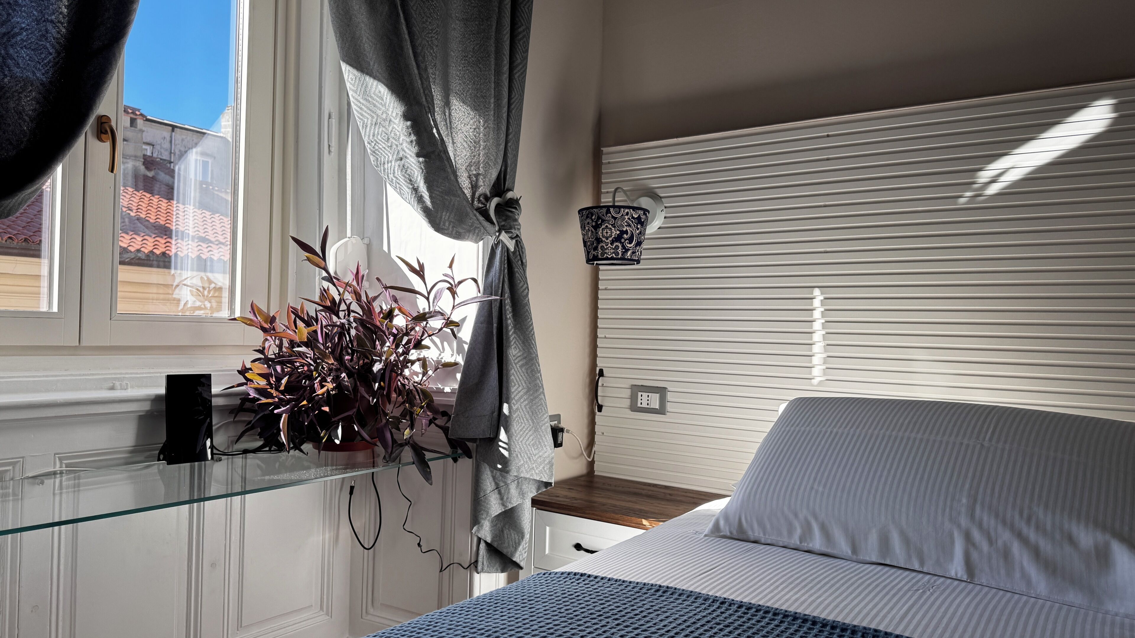 A bedroom with a bed, a window with curtains, a potted plant, and a wall-mounted lamp.