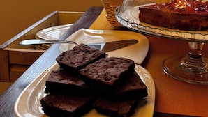 A table with a plate of brownies, a glass display case with a dessert, and a window view of buildings. - Bed & Chic art rooms (Trieste)