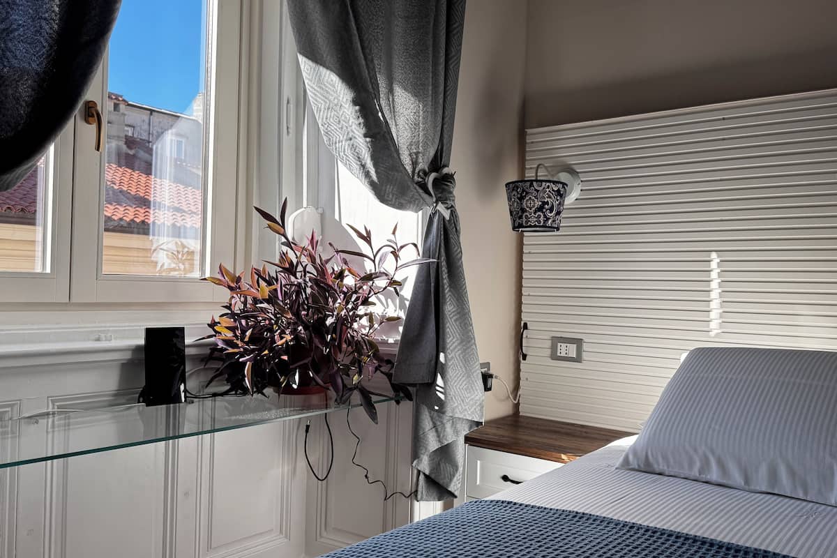 A bedroom with a bed, a window with curtains, a potted plant, and a wall-mounted lamp.