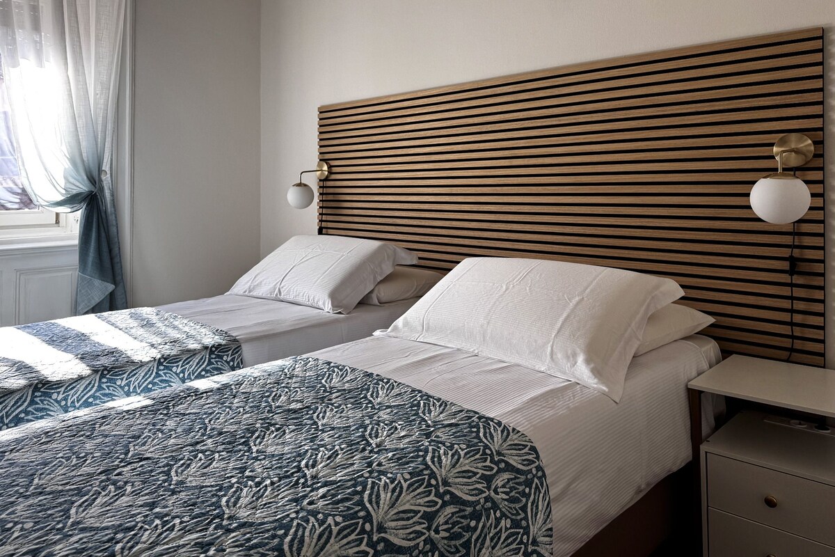 A hotel room with two beds, a wooden slatted headboard, bedside lamps, and a nightstand with drawers.