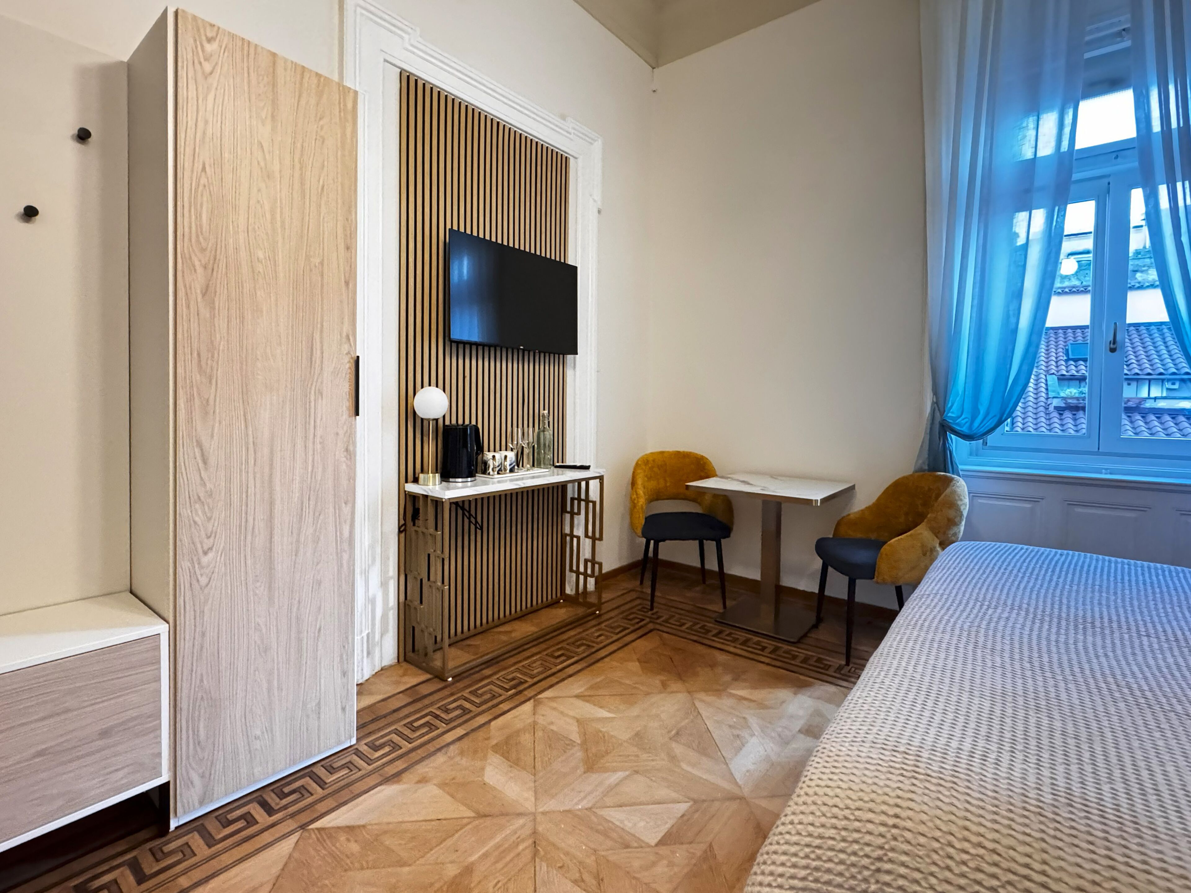 A hotel room with a bed, a TV mounted on a wooden panel wall, a small table with two chairs, and a window with blue curtains.
