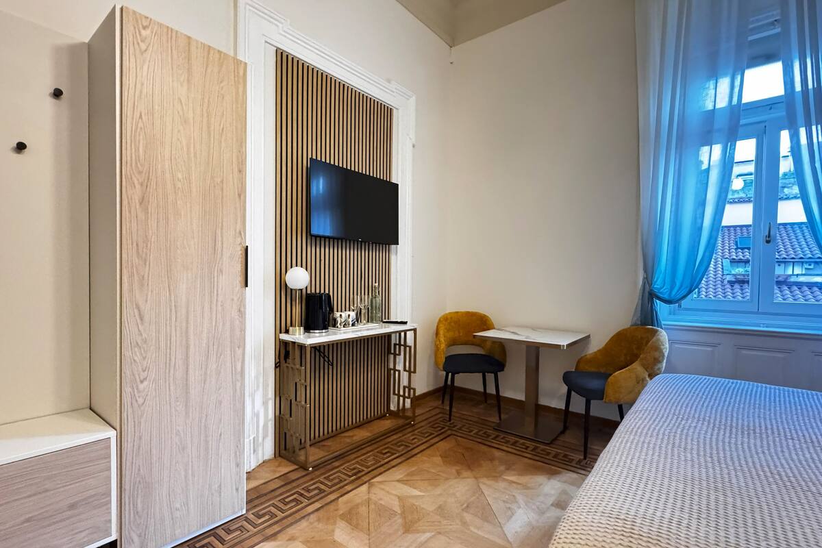 A modern hotel room with a bed, a TV mounted on a wooden panel wall, a small table with two chairs, and a window with blue curtains.