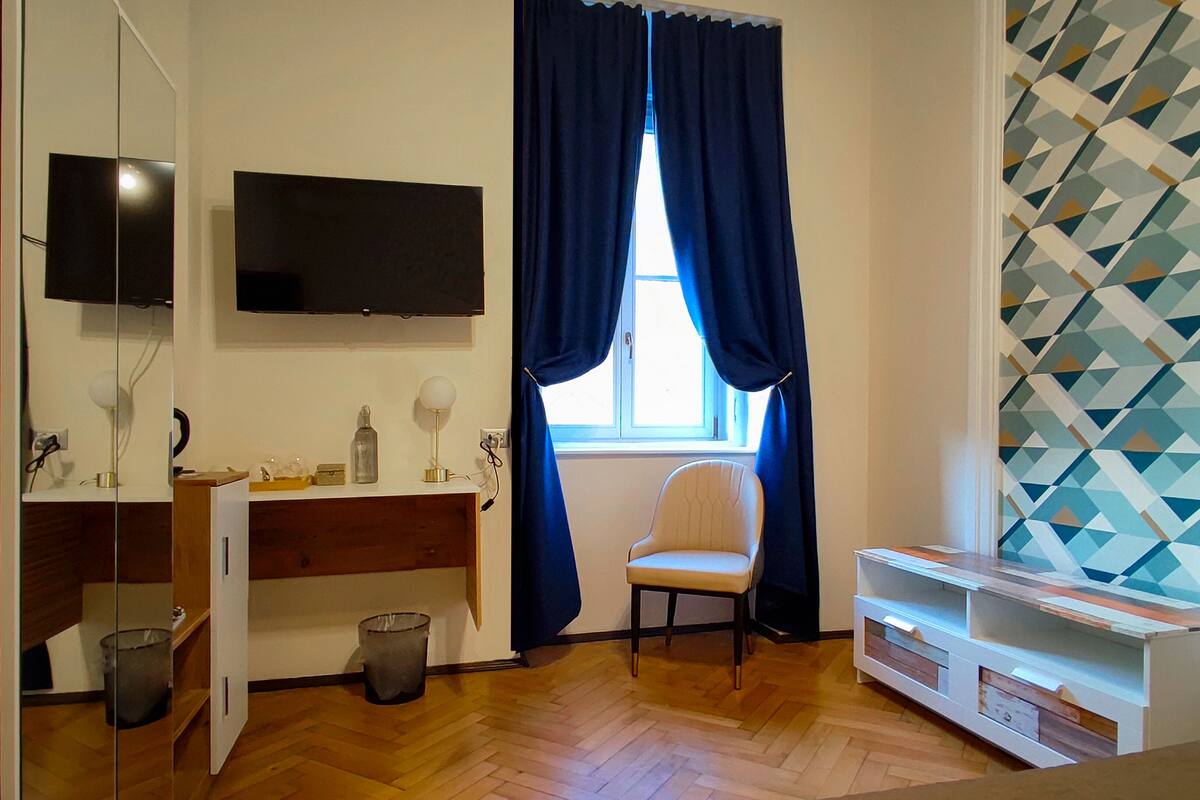 A room with a wooden floor, a white chair, a television mounted on the wall, a window with blue curtains, and a patterned wall.