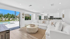 A modern living room with a white sofa, a round coffee table, and a view of a pool and outdoor area. - Coastal Getaway w/ Pool, Spa & Tiki Lounge (Naples)