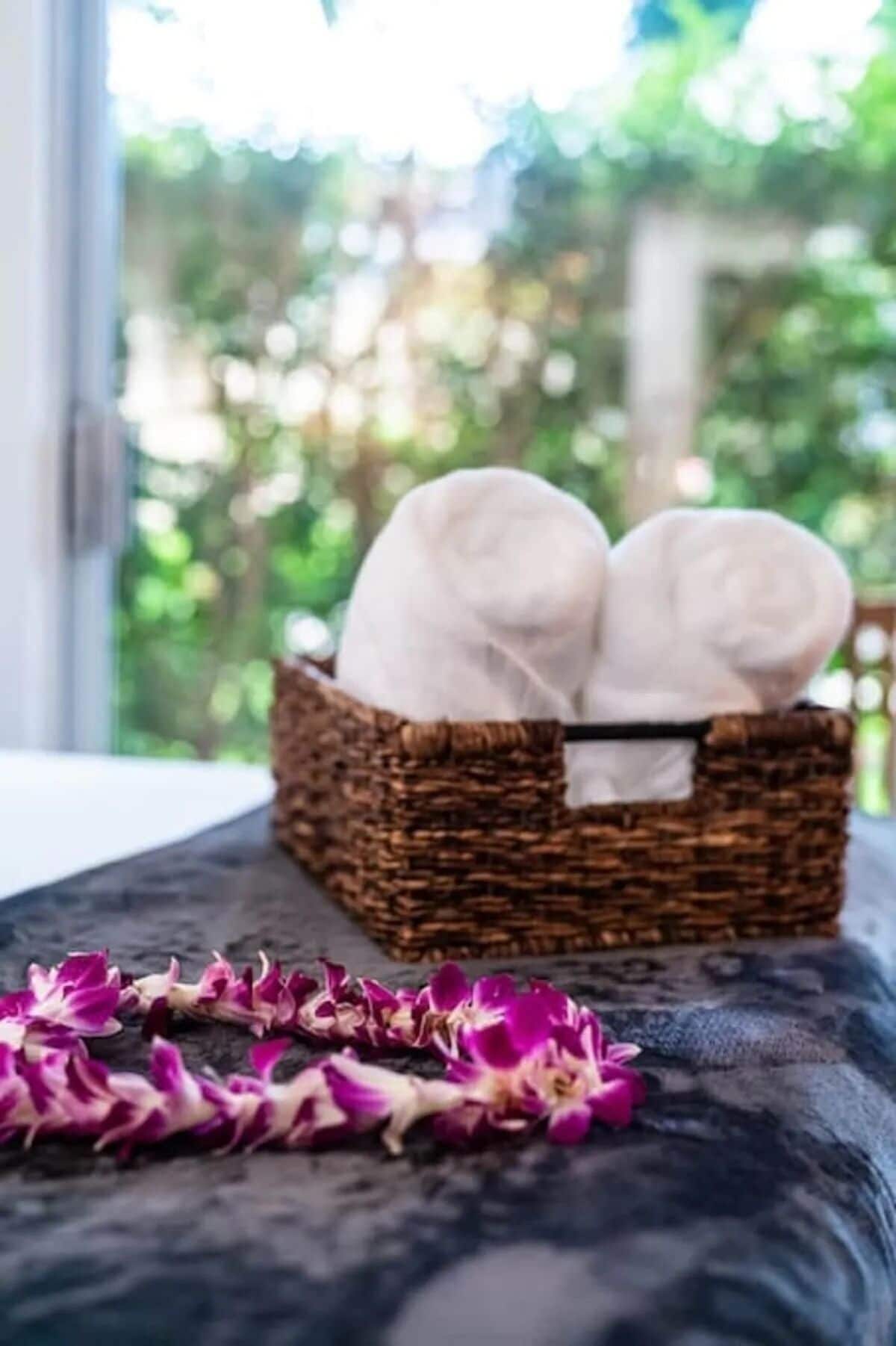 A wicker basket with two white towels on a dark-colored sofa near a window with a view of greenery.