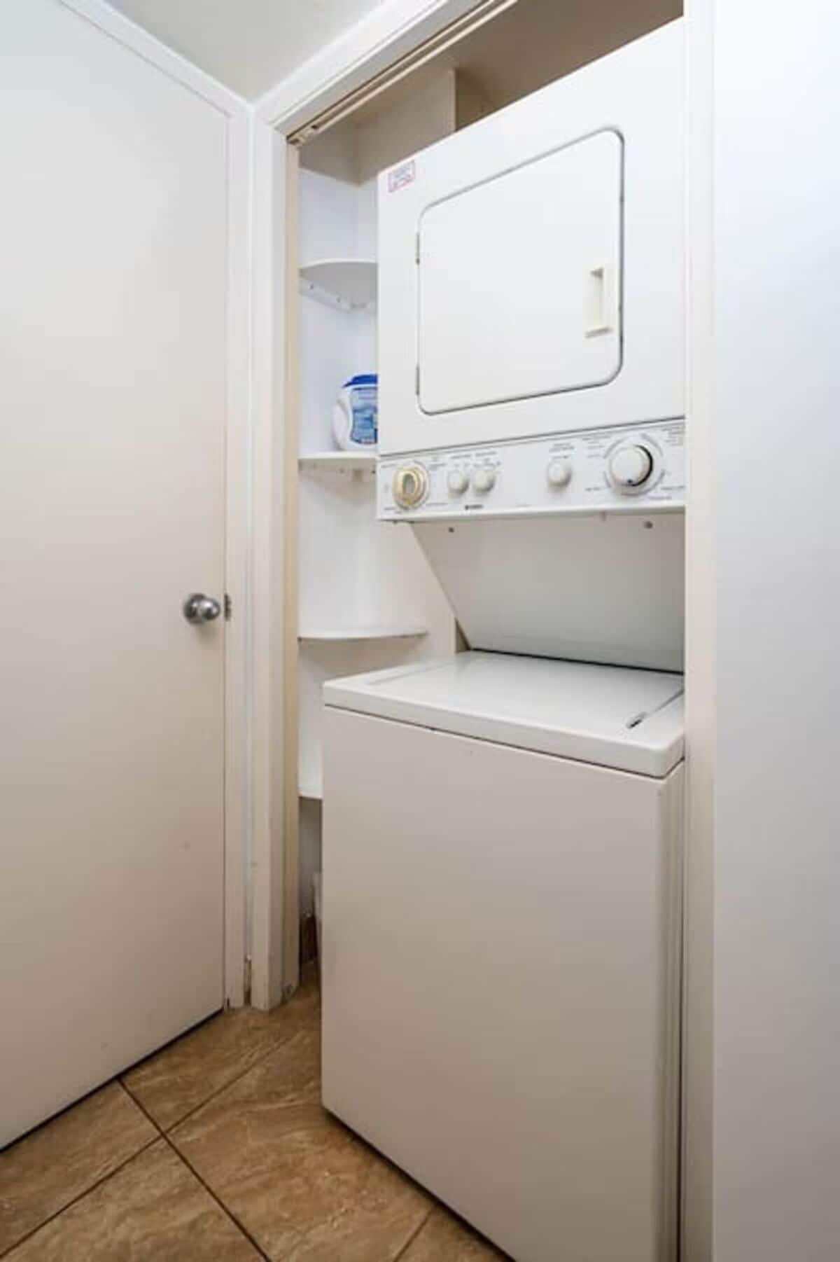 A compact laundry room with a front-loading washing machine and a dryer, a small shelf with a container, and a tiled floor.