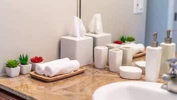 Un baño con encimera de mármol, lavamanos blanco, espejo, caja de pañuelos, dispensador de jabón y pequeñas plantas en macetas.