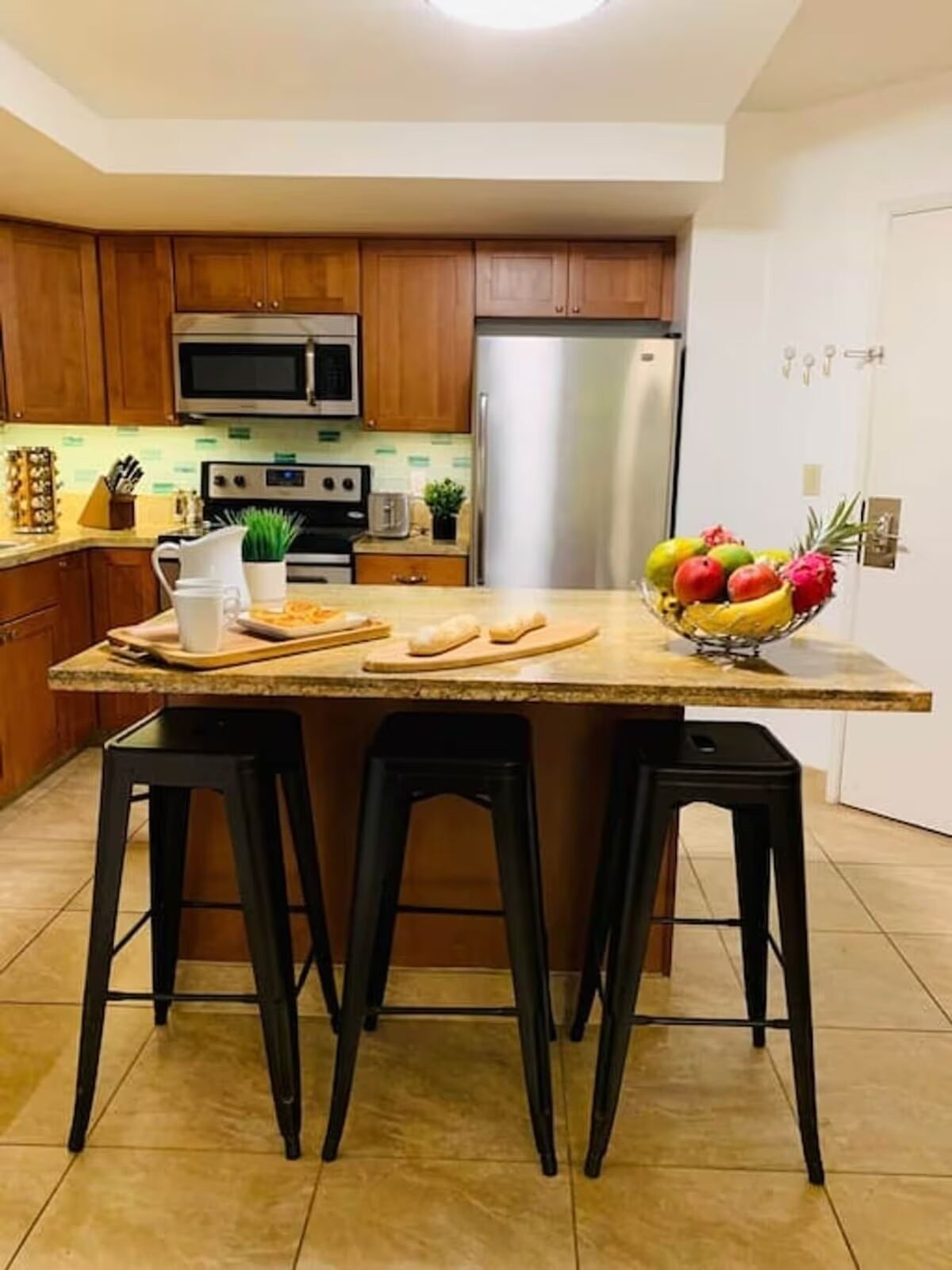 A kitchen with wooden cabinets, a stainless steel refrigerator, a microwave, a toaster, and a fruit bowl on the island. There are three black stools around the island.