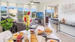 A breakfast table with various fruits, pastries, and beverages in a bright room with a view of the sea.