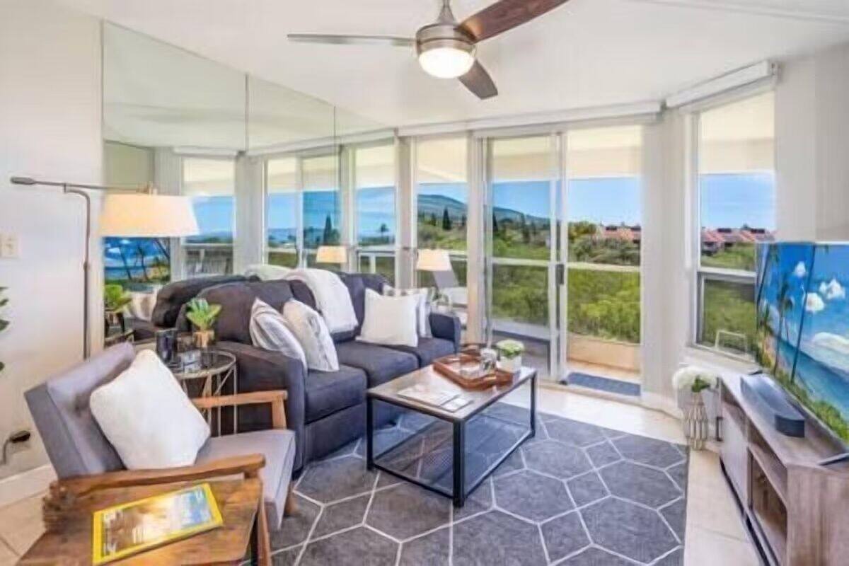 Un salon moderne avec un canapé, une table basse et un téléviseur, de grandes fenêtres offrant une vue sur le paysage et un ventilateur de plafond.