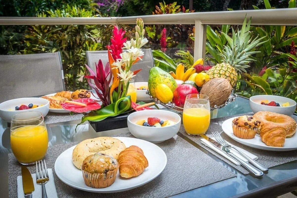 A breakfast table with various fruits, pastries, and a glass of orange juice on a balcony with a view of tropical plants.