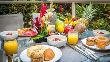 Una mesa de desayuno con diversas frutas, pasteles y un vaso de jugo de naranja en un balcón con vistas a plantas tropicales.
