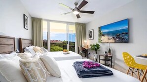 A hotel room with two beds, a ceiling fan, a large window with a view of the beach, a desk with a chair, and a TV on the wall.