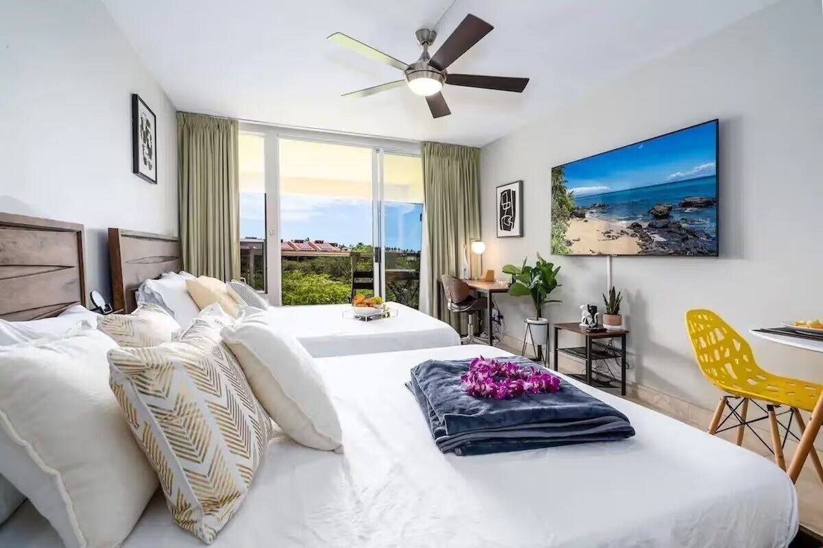 Une chambre d’hôtel avec deux lits, un ventilateur de plafond, une grande fenêtre donnant sur la plage, un bureau avec une chaise et un téléviseur fixé au mur.