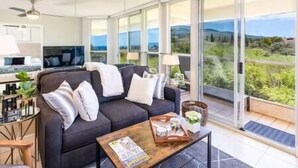 A modern living room with a dark sofa, a coffee table, and a view of a green landscape through large windows.