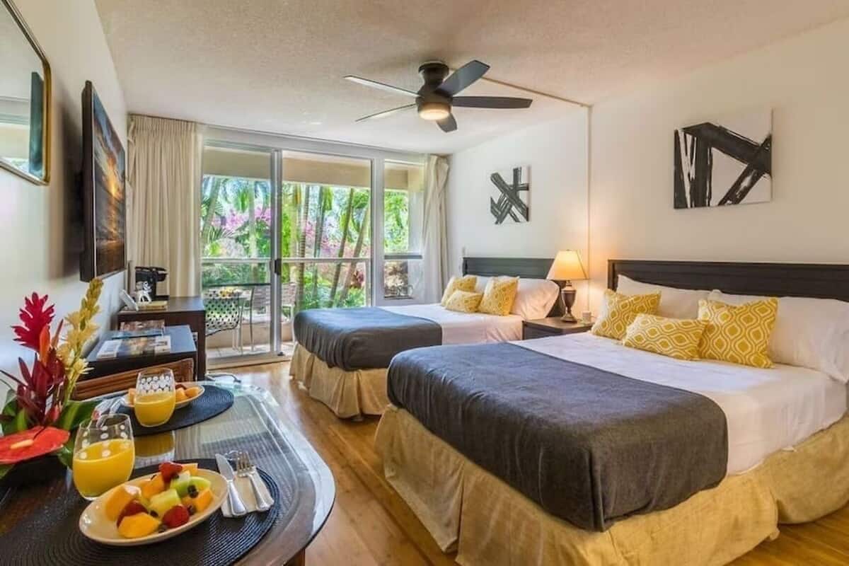 Une chambre d’hôtel avec deux lits, une table à manger où le petit-déjeuner est servi, un ventilateur de plafond et un balcon avec vue sur la verdure.