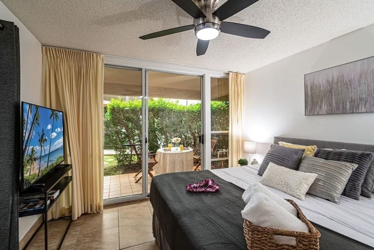 A bedroom with a bed, a ceiling fan, a TV, and a balcony with a table and chairs.
