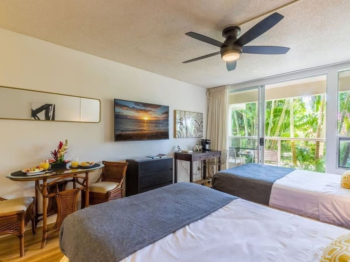 Ein Hotelzimmer mit zwei Betten, einem Schreibtisch, einem Stuhl, einem Deckenventilator und großen Fenstern mit Blick auf Grünflächen.