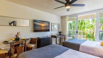 A hotel room with two beds, a desk, a chair, a ceiling fan, and large windows with a view of greenery.