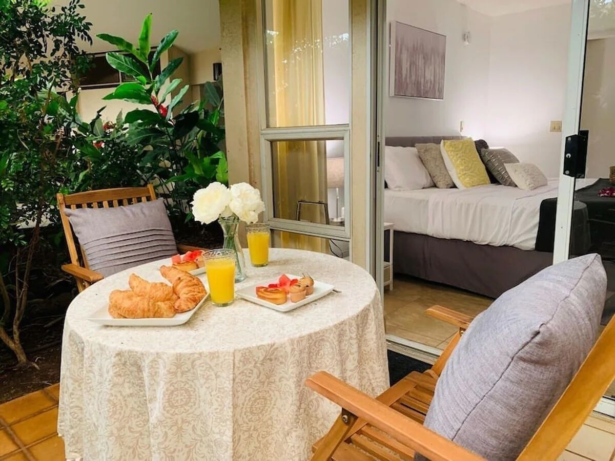 A dining area with a table set for breakfast, including croissants and juice, and a view of a bedroom with a bed and pillows.